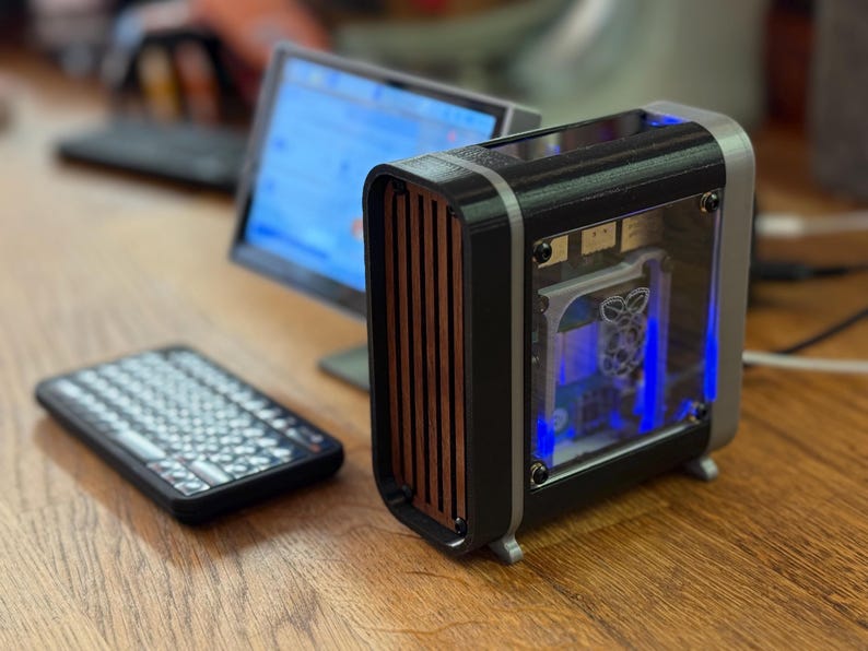 May include: A small, custom-built computer case with a black and silver frame, wood-grain front panel, and clear side panel. A small keyboard and monitor are also visible on a wooden desk. The case has blue internal lighting.