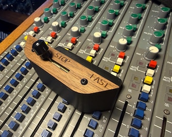 May include: A black and wood-grain controller featuring a sliding lever with a black knob. The wood surface is engraved with the text STOP on the left and FAST on the right. The device has a single black screw and is positioned on a mixing console.