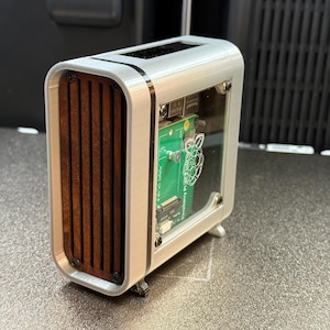May include: A silver-colored computer case with a clear front panel and wooden slats on the side. The case houses a green circuit board with a Raspberry Pi logo. The design is modern and compact, suitable for a small computer build.