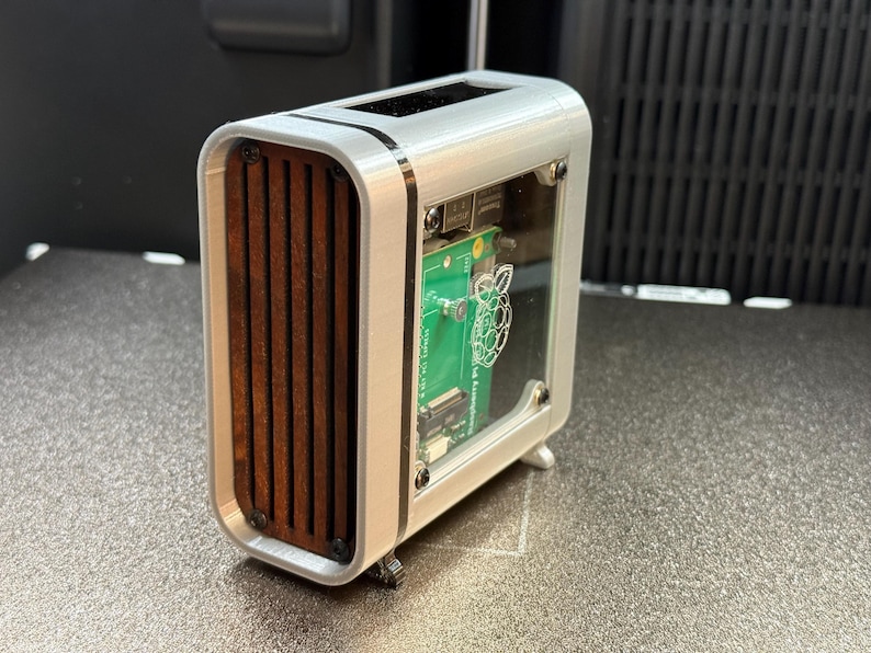 May include: A silver-colored computer case with a clear front panel and wooden slats on the side. The case houses a green circuit board with a Raspberry Pi logo. The design is modern and compact, suitable for a small computer build.