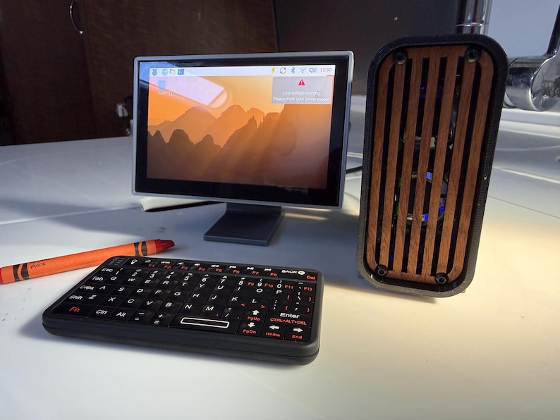 May include: A small computer setup featuring a monitor displaying a landscape image, a black keyboard, and a speaker with a wooden front. An orange crayon is also present. The monitor is approximately 6 inches wide.