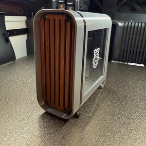 May include: A gray and wood-toned computer case with a clear window displaying a white Raspberry Pi logo. The front features vertical wooden slats. The case has a rounded, modern design.