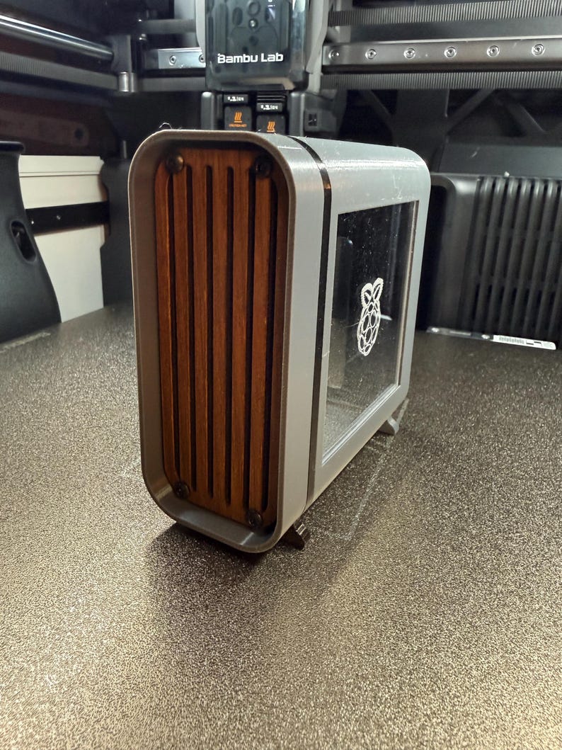 May include: A gray and wood-toned computer case with a clear window displaying a white Raspberry Pi logo. The front features vertical wooden slats. The case has a rounded, modern design.