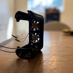 May include: Black electronic device with two small fans visible inside. The device has wires extending from it and is open, revealing the internal components. The device is sitting on a wooden surface.