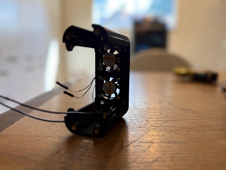 May include: Black electronic device with two small fans visible inside. The device has wires extending from it and is open, revealing the internal components. The device is sitting on a wooden surface.