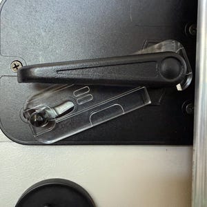 Puede incluir: Primer plano de un mecanismo de cierre negro con una palanca y componentes de plástico transparente. El pestillo está montado sobre una superficie gris oscuro, con una superficie blanca visible debajo. La imagen muestra los detalles del mecanismo de cierre.