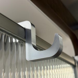 May include: A light gray, 3D-printed hook designed to hang over a horizontal bar. The hook has a curved shape and is mounted on a clear, textured surface. The hook is made of a light gray material.