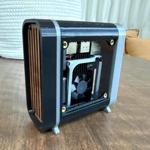 May include: A small, black and gray computer case with a fan visible inside. The case has a wooden front panel and is sitting on a wooden surface. The design is modern and compact.