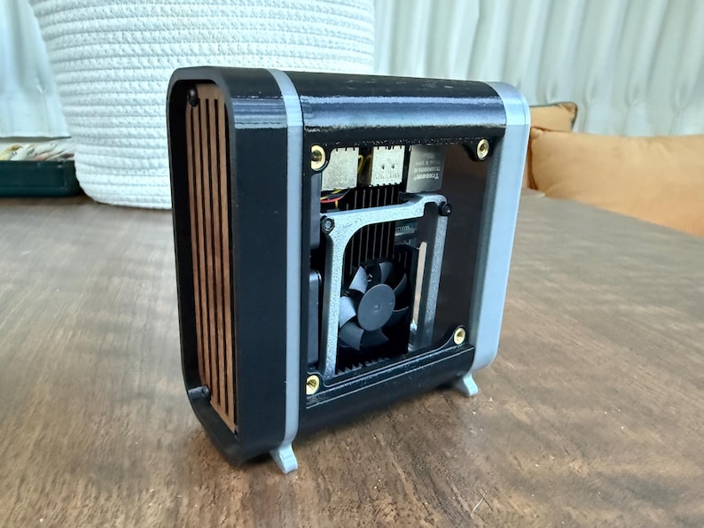 May include: A small, black and gray computer case with a fan visible inside. The case has a wooden front panel and is sitting on a wooden surface. The design is modern and compact.