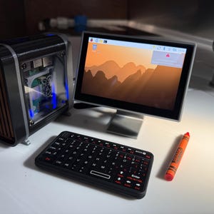 May include: A miniature computer setup featuring a black and brown computer case, a monitor displaying a mountain landscape, a black keyboard, and an orange crayon. The monitor is approximately 7 inches.