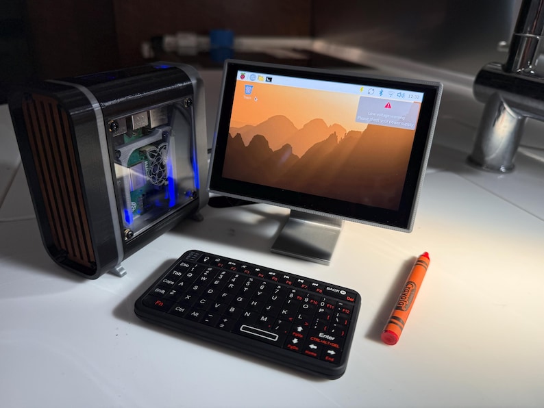 May include: A miniature computer setup featuring a black and brown computer case, a monitor displaying a mountain landscape, a black keyboard, and an orange crayon. The monitor is approximately 7 inches.
