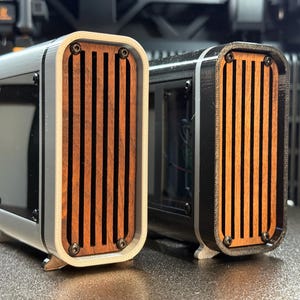 May include: Two rectangular speaker enclosures with wooden slats and black accents. One is white and the other is black. Both have a curved design and are mounted on small feet. The wooden slats are a warm brown color.