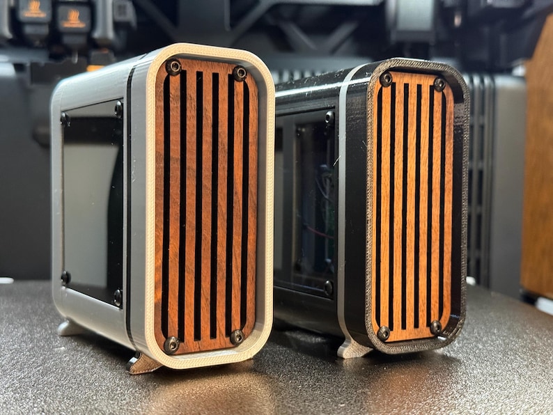 May include: Two rectangular speaker enclosures with wooden slats and black accents. One is white and the other is black. Both have a curved design and are mounted on small feet. The wooden slats are a warm brown color.
