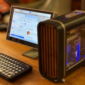 May include: A small computer setup featuring a monitor displaying a social media profile, a compact keyboard, and a desktop computer with a transparent side panel and wooden accents. The computer case has blue internal lighting.