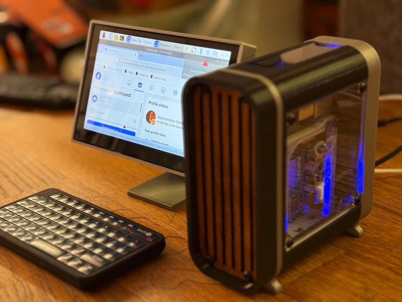 May include: A small computer setup featuring a monitor displaying a social media profile, a compact keyboard, and a desktop computer with a transparent side panel and wooden accents. The computer case has blue internal lighting.