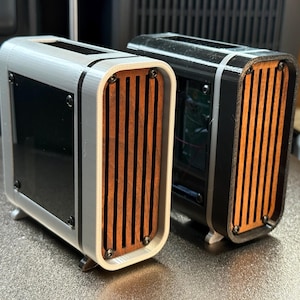 May include: Two small, rectangular computer cases. One is silver with black accents and a wooden slat front. The other is black with a wooden slat front. Both have small feet and a black glass panel.