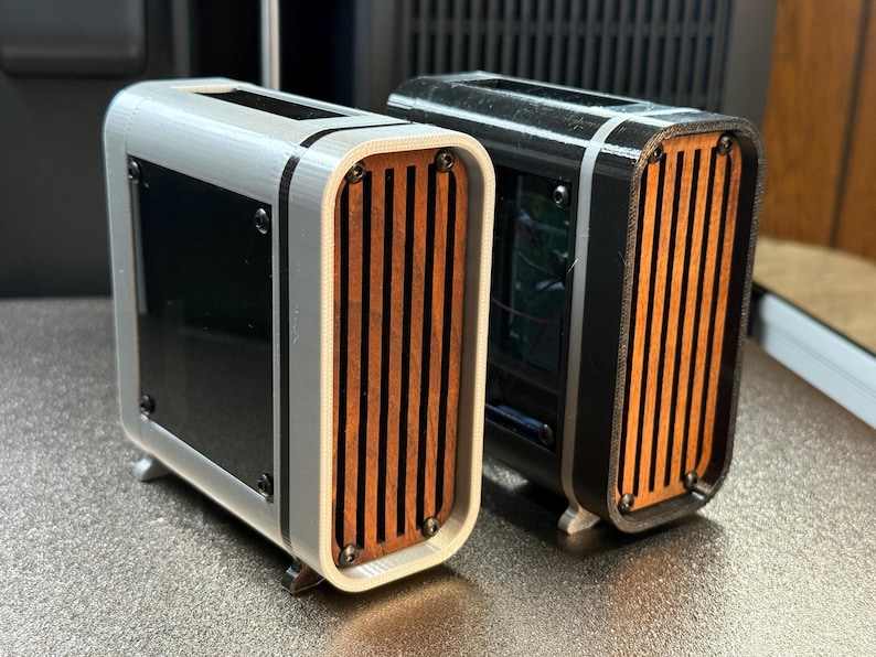 May include: Two small, rectangular computer cases. One is silver with black accents and a wooden slat front. The other is black with a wooden slat front. Both have small feet and a black glass panel.