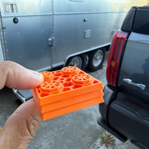 May include: A stack of four bright orange, square-shaped building blocks. Each block has a grid pattern and four circular designs on the top surface. The blocks are held in front of a silver trailer and a dark gray vehicle.