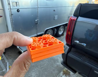 May include: A stack of four bright orange, square-shaped building blocks. Each block has a grid pattern and four circular designs on the top surface. The blocks are held in front of a silver trailer and a dark gray vehicle.