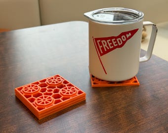 May include: A white mug with a handle and a lid, featuring a red flag design with the word "FREEDOM". The mug sits on an orange coaster, next to a stack of similar coasters. The coasters have a geometric design.