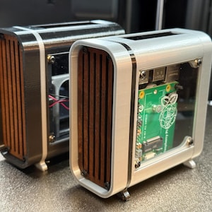 May include: Two small, rectangular computer cases. One is black with a silver trim, the other is silver with a white trim. Both have wooden slats and a clear front panel revealing a green circuit board. The cases have small feet.