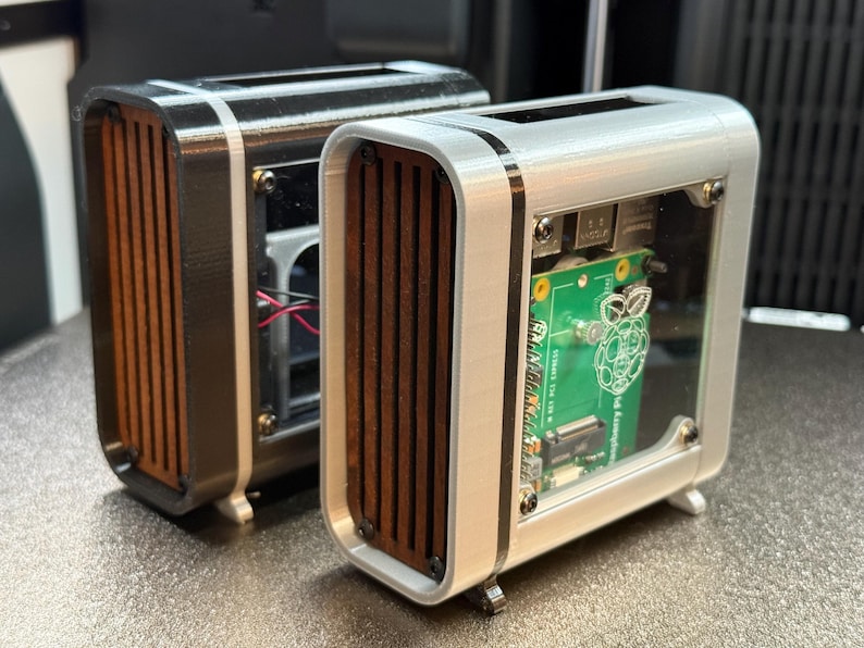 May include: Two small, rectangular computer cases. One is black with a silver trim, the other is silver with a white trim. Both have wooden slats and a clear front panel revealing a green circuit board. The cases have small feet.