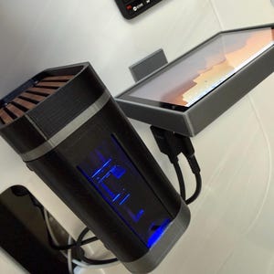 May include: A black and gray electronic device with a blue illuminated display, a tablet, and a small black keyboard. The device has a rectangular shape with a silver trim and a wooden top. The tablet is in a gray frame.