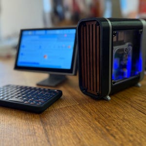 May include: A small computer setup featuring a black keyboard, a monitor displaying a blue screen, and a compact black and wood-toned computer case. The case has a clear side panel, revealing internal components.