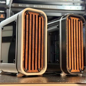 May include: Two rectangular enclosures with wooden slat fronts. One is white and the other is black. Both have rounded corners and are mounted on small feet. The wooden slats are a warm brown color.