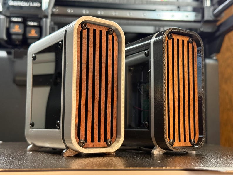 May include: Two rectangular enclosures with wooden slat fronts. One is white and the other is black. Both have rounded corners and are mounted on small feet. The wooden slats are a warm brown color.