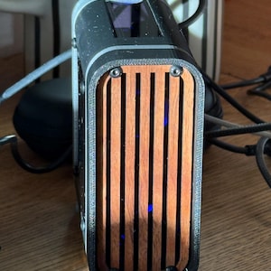 May include: A black and wood-toned electronic device with a rectangular shape. The front features vertical wooden slats, and the top has a dark screen. The device is secured with black screws and has a sleek, modern design.