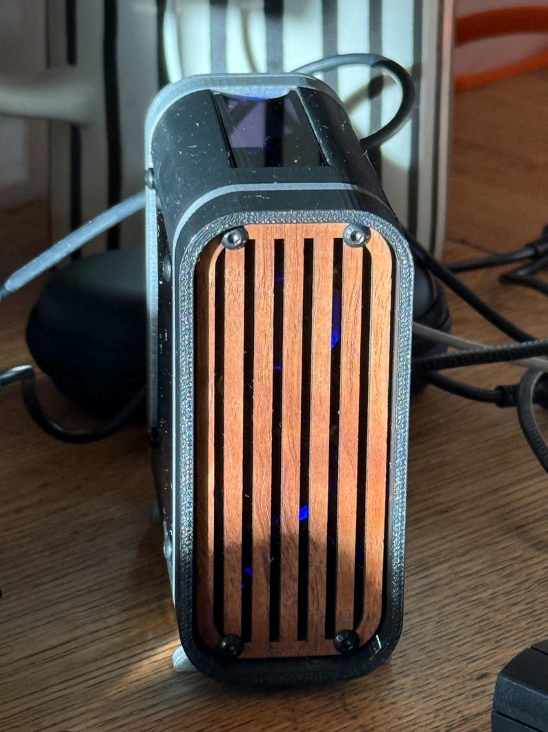 May include: A black and wood-toned electronic device with a rectangular shape. The front features vertical wooden slats, and the top has a dark screen. The device is secured with black screws and has a sleek, modern design.