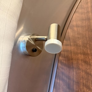 May include: A brushed nickel-finished wall-mounted light fixture with a cylindrical head and a white, circular light diffuser. The fixture is mounted on a metallic surface, with a wooden panel visible in the background.