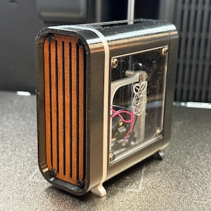 May include: A small, rectangular black and silver electronic device with a wooden slat front. The device has a clear panel revealing internal components and wiring. The design is modern and compact, with a focus on contrasting materials.