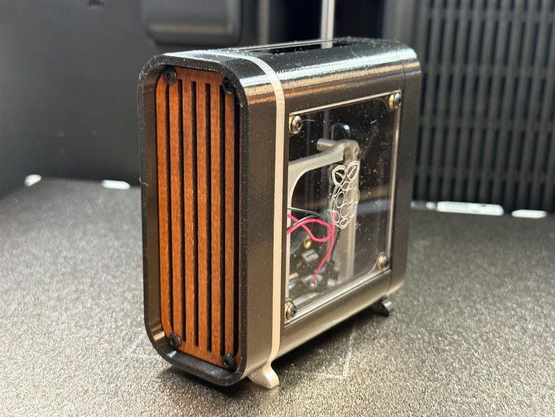 May include: A small, rectangular black and silver electronic device with a wooden slat front. The device has a clear panel revealing internal components and wiring. The design is modern and compact, with a focus on contrasting materials.