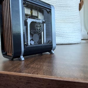 May include: A black and silver computer case with a clear front panel, revealing internal components including a fan and circuit board. The side panel features vertical wooden slats. The case sits on a wooden surface.