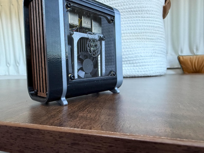 May include: A black and silver computer case with a clear front panel, revealing internal components including a fan and circuit board. The side panel features vertical wooden slats. The case sits on a wooden surface.