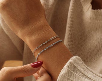 Puede incluir: Dos delicadas pulseras de plata con pequeñas piedras redondas y transparentes se llevan en la muñeca. Las pulseras están hechas de una fina cadena con piedras espaciadas uniformemente. Se ve la mano de la persona, con esmalte de uñas rojo.