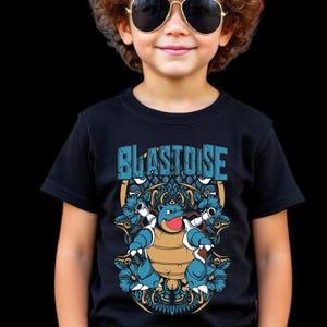 Puede incluir: Una camiseta negra con un diseño de dibujos animados de Blastoise y la palabra "BLASTOISE" en azul. El diseño incluye elementos florales azules y dorados. La persona de la imagen lleva gafas de sol de aviador con montura dorada.