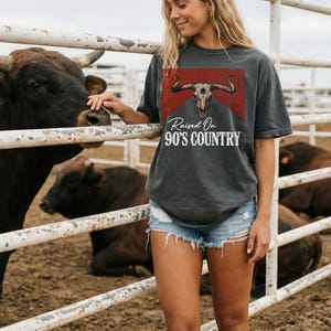 May include: Dark gray t-shirt with a graphic of a bull skull on a red background, with the text "Raised On 90's Country" in white. The shirt is paired with denim shorts. The image is taken outdoors near a metal fence.