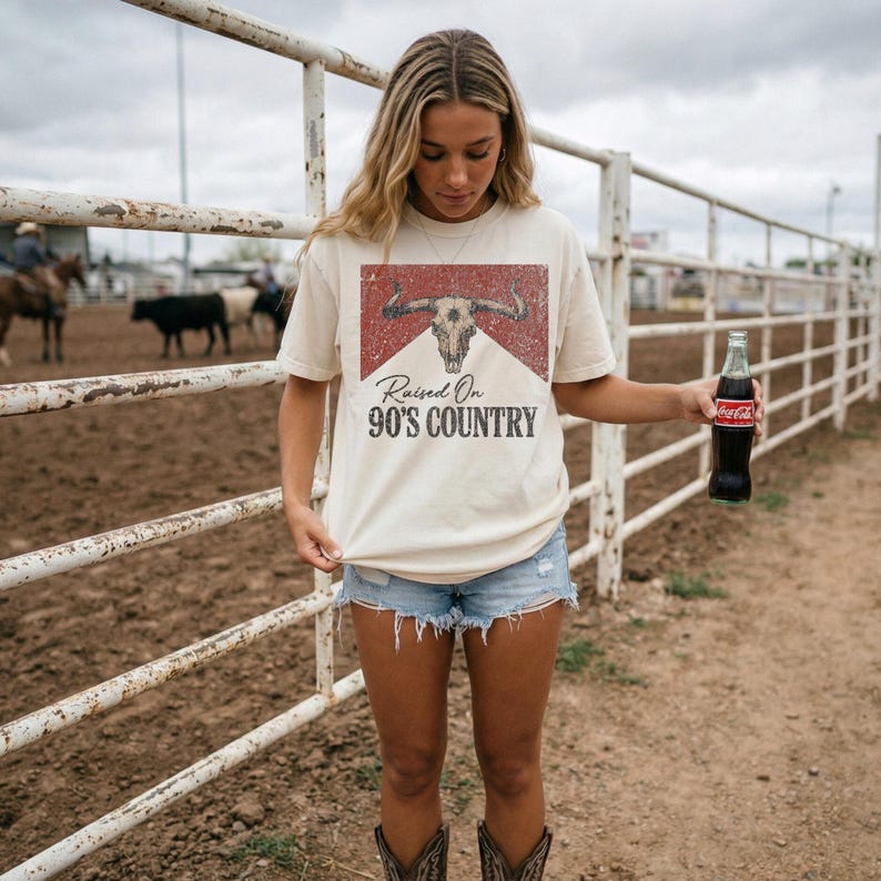 May include: Cream-colored t-shirt with a distressed graphic of a bull skull and the text "Raised On 90's Country." The shirt is paired with denim shorts and cowboy boots. A glass bottle of Coca-Cola is held in the hand.