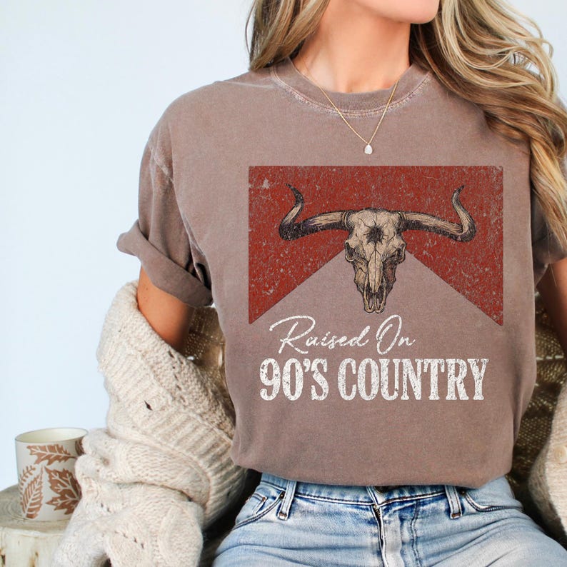 May include: A brown t-shirt with a graphic of a bull skull and the text "Raised On 90's Country". The design is set against a red background. The shirt has rolled-up sleeves and a casual fit.