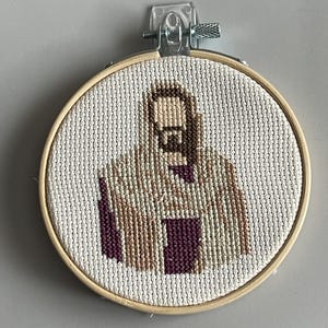 Puede incluir: Un bastidor de bordado circular de madera que contiene un dise&ntilde;o de punto de cruz sobre tela Aida blanca. El dise&ntilde;o representa una figura estilizada con una t&uacute;nica con capucha en tonos beige, marr&oacute;n y morado oscuro, centrada en el bastidor.