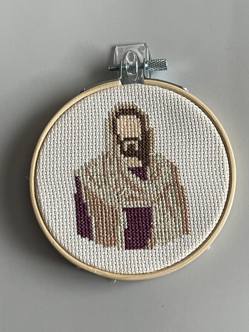 Puede incluir: Un bastidor de bordado circular de madera que contiene un dise&ntilde;o de punto de cruz sobre tela Aida blanca. El dise&ntilde;o representa una figura estilizada con una t&uacute;nica con capucha en tonos beige, marr&oacute;n y morado oscuro, centrada en el bastidor.