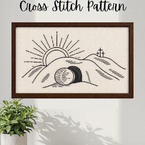 Op de afbeelding: Een ingelijst kruissteekpatroon met een zonsopgang boven heuvels, drie kruisen en een graf. De tekst "Cross Stitch Pattern" staat boven de afbeelding. Het kunstwerk is in zwart en wit.