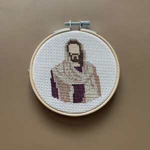 Puede incluir: Un bastidor de bordado circular de madera que contiene un patr&oacute;n de punto de cruz sobre tela Aida blanca. El dise&ntilde;o representa una figura con barba y cabello casta&ntilde;os, vistiendo una t&uacute;nica color canela con una prenda morada oscura debajo.