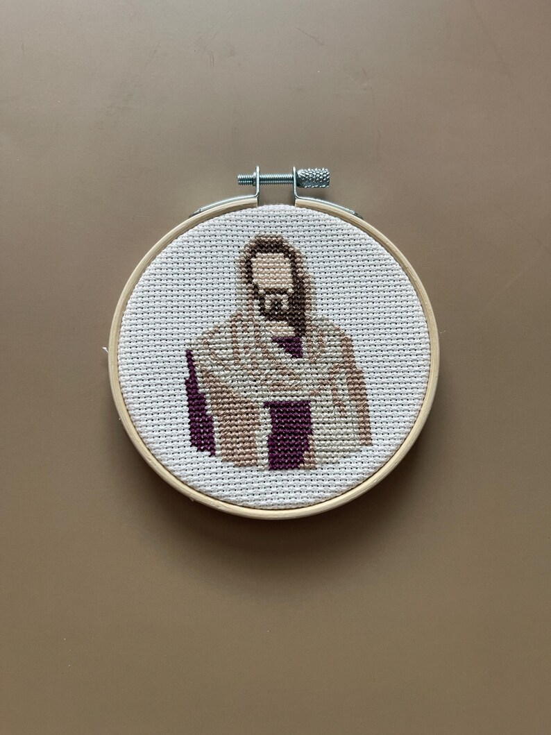 Puede incluir: Un bastidor de bordado circular de madera que contiene un patr&oacute;n de punto de cruz sobre tela Aida blanca. El dise&ntilde;o representa una figura con barba y cabello casta&ntilde;os, vistiendo una t&uacute;nica color canela con una prenda morada oscura debajo.
