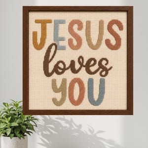 Op de afbeelding: Kruissteekpatroon met de woorden "JESUS loves YOU" in verschillende kleuren, omlijst in een donkerbruine houten lijst. De tekst is geborduurd op een beige achtergrond. De woorden "Cross Stitch Pattern" staan bovenaan.