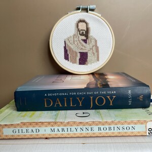 Puede incluir: Un bastidor de bordado circular de madera que contiene un patr&oacute;n de punto de cruz de una figura con barba que lleva una t&uacute;nica color canela sobre una prenda morada. El bastidor est&aacute; colocado detr&aacute;s de una pila de tres libros titulados Daily Joy, Gilead y The Holy Temple.