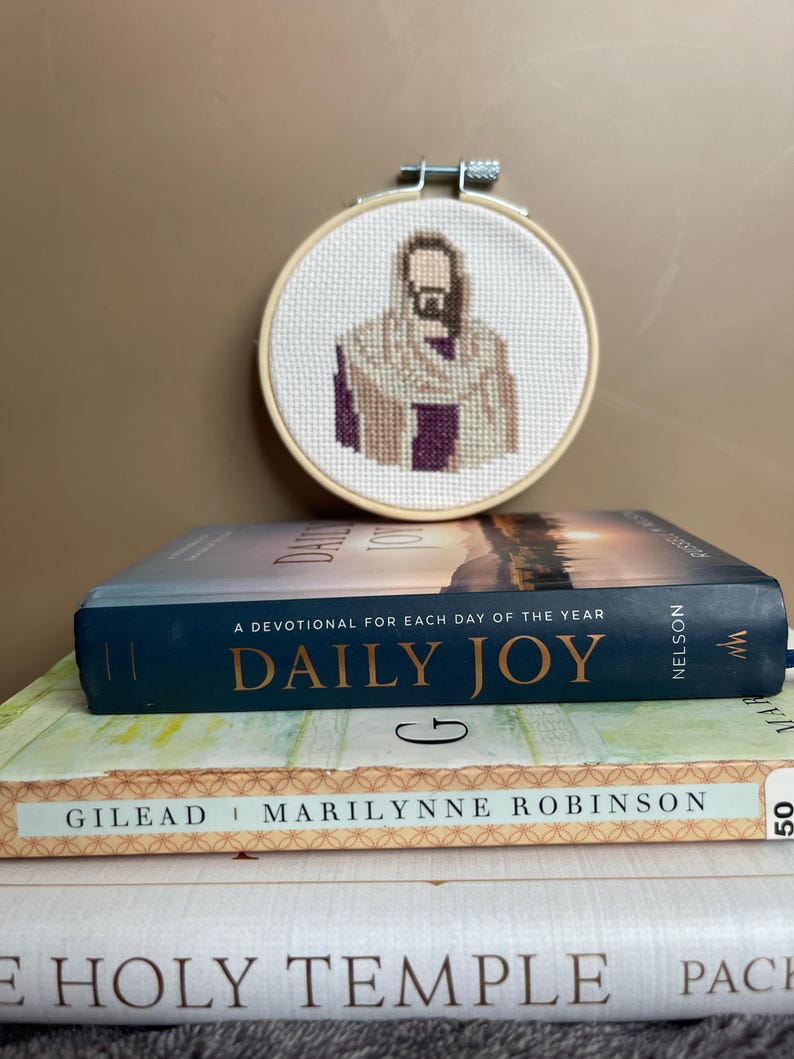 Puede incluir: Un bastidor de bordado circular de madera que contiene un patr&oacute;n de punto de cruz de una figura con barba que lleva una t&uacute;nica color canela sobre una prenda morada. El bastidor est&aacute; colocado detr&aacute;s de una pila de tres libros titulados Daily Joy, Gilead y The Holy Temple.